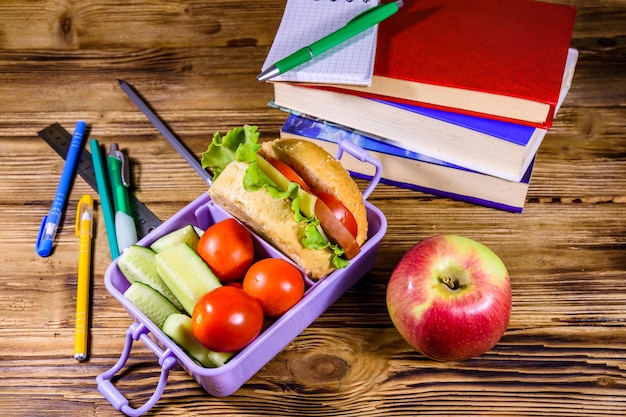 Canetas de bloco de notas pilha de livros e lancheira com pepinos de hambúrguer e tomate na mesa de madeira rústica