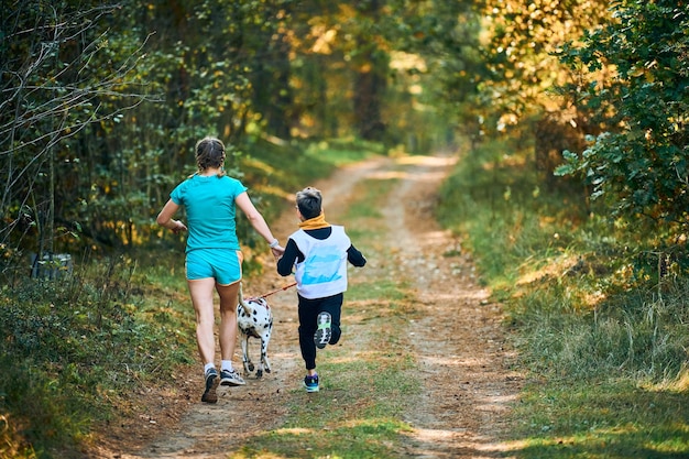 Canicross-Hunde-Mushing-Rennen, dalmatinischer Hund, der an der Mutter und ihrem Sohn befestigt ist, Joggen entlang der Rückansicht des Waldweges, Hunderennen, aktives Familienjoggen im Freien