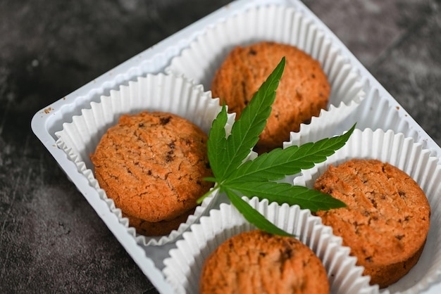 Cannabis Food Cookies Box mit Cannabisblatt Marihuana-Kraut auf dunklem Hintergrund, köstlicher süßer Dessert-Cookie mit Hanfblattpflanze THC CBD-Kräuter-Food-Snack und medizinisches Konzept