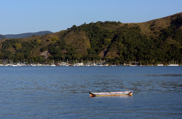 Canoa solitária em mar calmo, com barcos, morro e céu azul