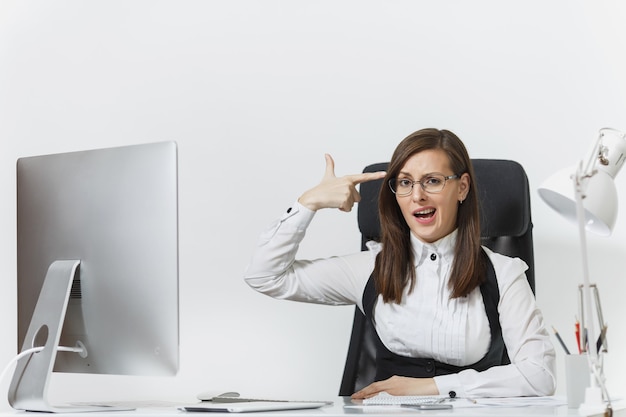 Cansada, perplexa e estressada, mulher de negócios de terno sentada à mesa, trabalhando em um computador contemporâneo com documentos, colocando a mão na cabeça como uma pistola para atirar no escritório