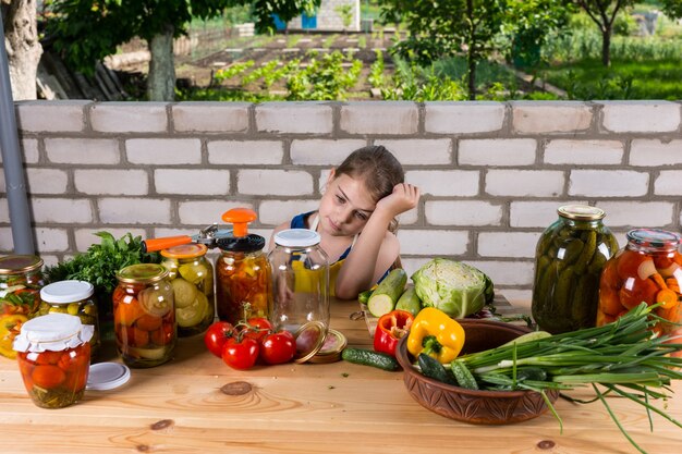 Cansado buscando joven apoyado en la mano, sentado en la mesa cubierta con verduras frescas y frascos de conservas en escabeche al aire libre en el jardín