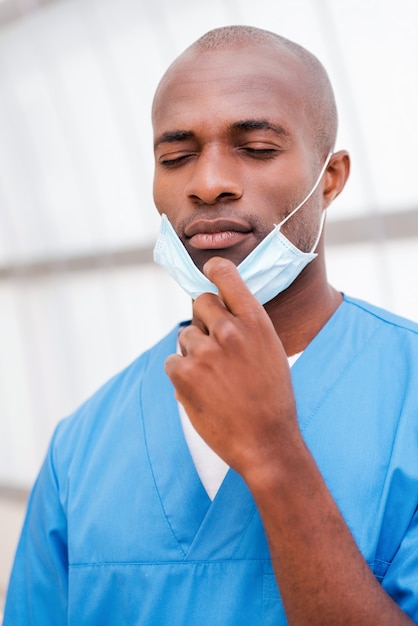 Cansado después de una larga cirugía. Retrato de joven médico africano cansado en uniforme azul tomando su mascarilla quirúrgica y manteniendo los ojos cerrados