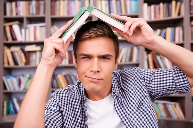 Cansado de estudiar. Hombre joven frustrado que lleva un libro en la cabeza y que expresa negatividad mientras está sentado contra la estantería