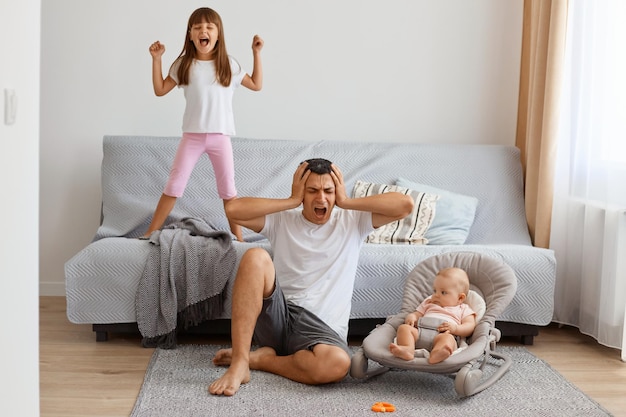 Cansado gritando guapo padre morena pasar tiempo con hijas mientras está sentado en el piso cerca del sofá, tapándose los oídos con las manos, no quiere escuchar a la hija mayor gritando.