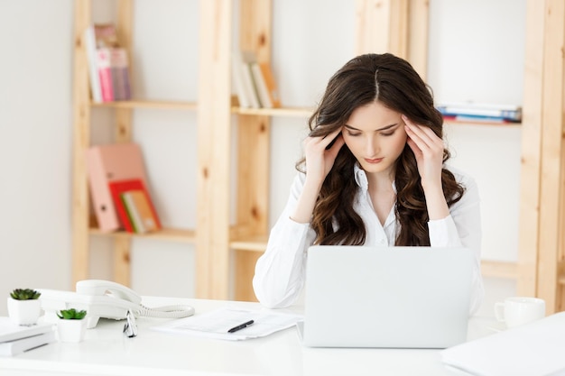 Cansado hermosa empresaria sosteniendo la mano en la cabeza mientras trabaja en la computadora y algunos documentos comerciales en la oficina brillante