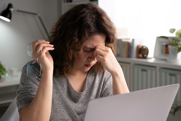 Foto cansado jovem asiático com dor nos olhos durante o trabalho em home office usando laptop. aluna asiática cansada e sobrecarregada segurando óculos sente fadiga ocular após o trabalho no computador