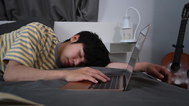 Cansado jovem asiático dormindo na cama perto do computador portátil