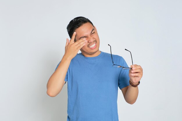 Cansado jovem asiático em camiseta azul tirando óculos sofrendo de enxaqueca ou dor de cabeça isolada no fundo branco