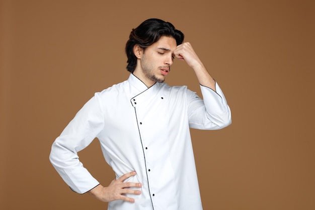 Cansado jovem chef masculino vestindo uniforme, mantendo a mão na cintura tocando a testa com os olhos fechados, isolados no fundo marrom