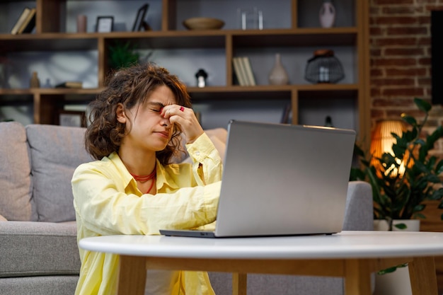 Cansado joven trabajador estudiante rizado hace una pausa en el trabajo en línea se quita las gafas masajea el puente de la nariz Burnout fatiga exceso de trabajo en la computadora fatiga que sufre de fatiga visual