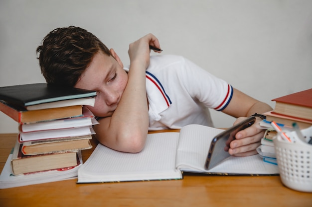 Foto cansado de lecciones, un adolescente en casa para libros de texto mira un teléfono inteligente