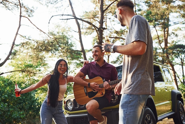 Cantando canções juntos. amigos têm um bom fim de semana ao ar livre perto de seu carro verde com violão.