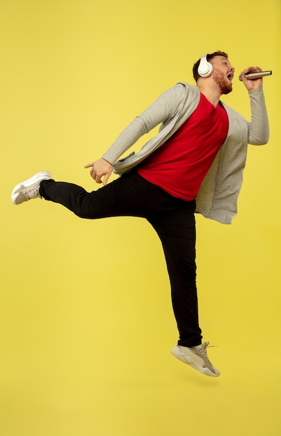 Foto cantando com fones de ouvido. retrato de corpo inteiro de jovem salto em altura isolado no fundo amarelo do estúdio. modelo masculino caucasiano atraente. copyspace. emoções humanas, conceito de expressão facial.