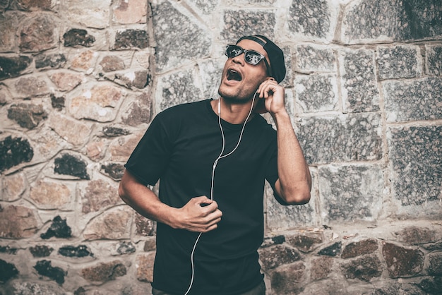 Cantando como uma estrela. Africano jovem bonito com fones de ouvido cantando e gesticulando em pé contra a parede de pedra ao ar livre