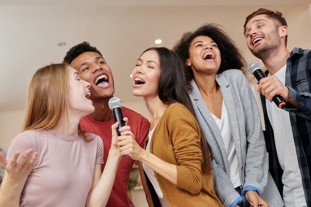 Cantando para todos, amigos multiculturais alegres cantando com microfone enquanto jogam karaokê