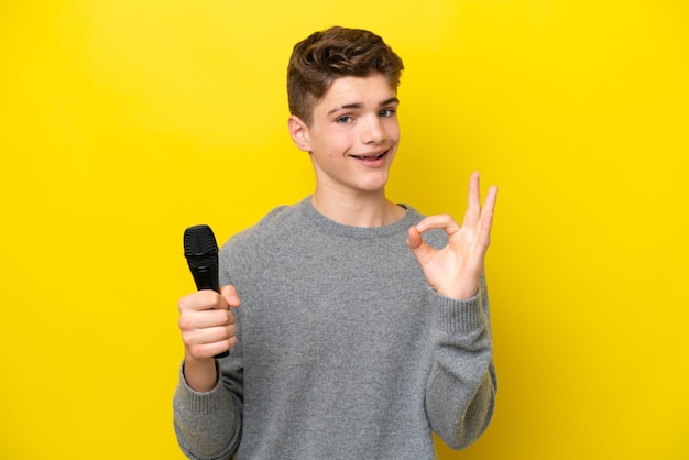Cantante Adolescente hombre recogiendo un micrófono aislado sobre fondo amarillo que muestra el signo de ok con los dedos