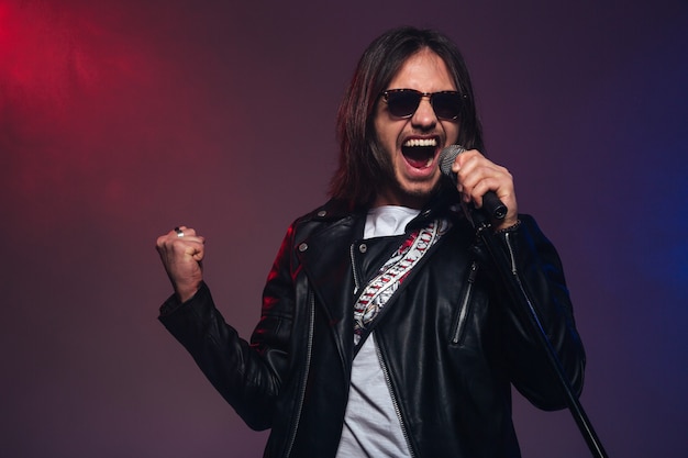 Foto cantante masculino joven atractivo en gafas de sol con pelo largo cantando con micrófono sobre fondo ahumado colorido