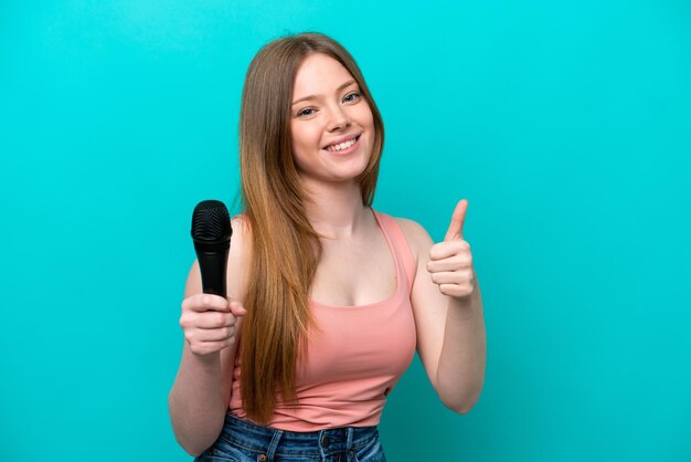 Cantante mujer caucásica recogiendo un micrófono aislado de fondo azul con los pulgares hacia arriba porque algo bueno ha sucedido