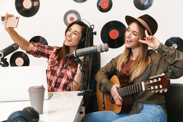 Cantante de mujer feliz actuando en un programa de radio mientras graba un podcast para un programa en línea, toca la guitarra, se toma una selfie con un anfitrión