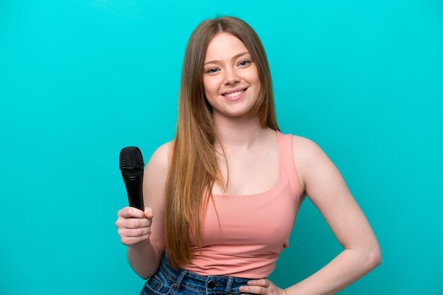 Cantora mulher caucasiana pegando um microfone isolado em fundo azul posando com braços no quadril e sorrindo