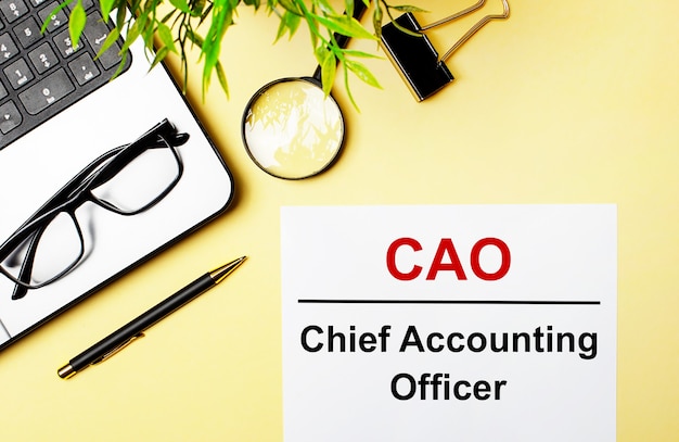 CAO Chief Accounting Officer steht in Rot auf einem weißen Blatt Papier auf hellgelbem Hintergrund neben einem Laptop, einem Stift, einer Lupe, einer Brille und einer grünen Pflanze.