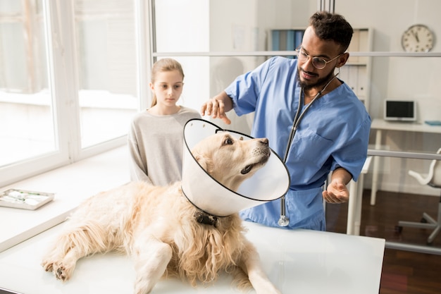 Cão doente com funil no pescoço, deitado na mesa e olhando para o jovem médico veterinário profissional