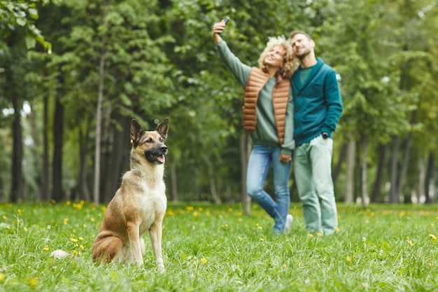 Cão pastor sentado na grama verde com o jovem casal fazendo selfie ao ar livre