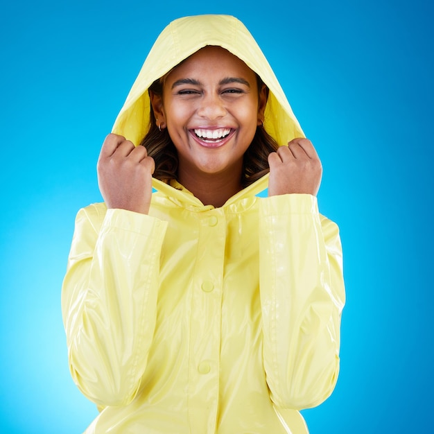 Foto capa feliz e retrato de uma mulher com uma capa de chuva isolada em um fundo azul em um estúdio sorria rindo e menina vestindo uma jaqueta para proteger da chuva o frio ou o mau tempo em um pano de fundo