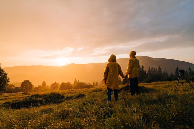 capas de chuva amarelas ficam em um prado nas montanhas e assistem o belo pôr do sol