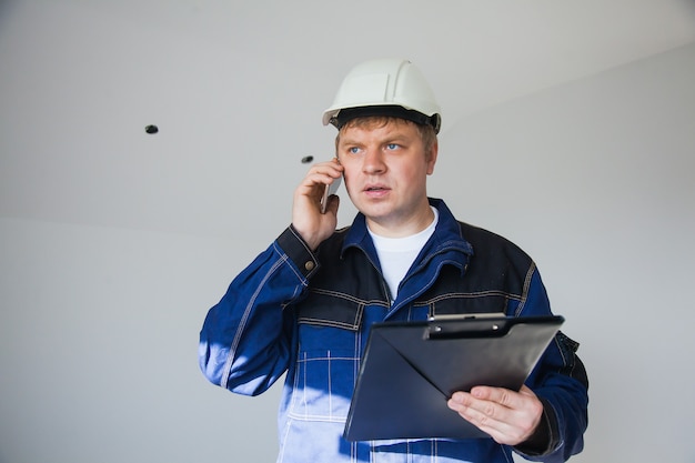 Capataz de constructor en un casco blanco en un sitio de construcción en el interior, concepto de renovación plana, capataz profesional en el trabajo con smartphone