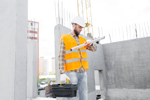 El capataz del constructor con casco y chaleco de protección está de pie en el lugar de trabajo en el edificio y sostiene la regla y la caja de herramientas en las manos