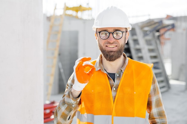 El capataz del constructor con casco protector y chaleco se encuentra en su lugar de trabajo en el edificio en construcción y muestra un gesto de aprobación