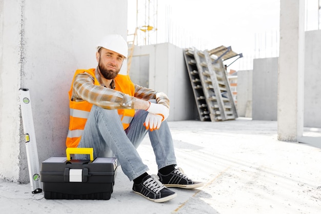 Capataz de reparador de construtor em capacete de segurança e colete sentado para descansar no local de trabalho na construção ao lado de caixa de ferramentas e régua