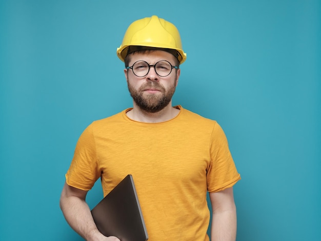 Capataz o ingeniero masculino sospechoso en un casco de construcción y soportes de anteojos redondos con una computadora portátil