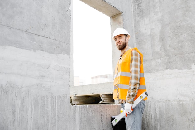 Capataz de reparador de constructores en casco protector y chaleco se para en el lugar de trabajo en el edificio y sostiene la regla y la caja de herramientas en las manos