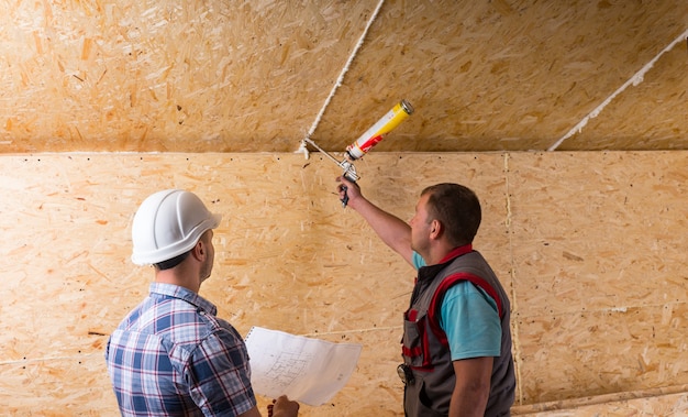 Capataz del sitio de construcción con casco blanco sosteniendo planes y trabajador de observación que aplica calafateo al techo de madera sin terminar