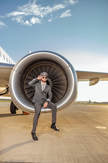 Capitán de la aerolínea barbudo alegre ajustando gafas de sol y sonriendo mientras está sentado en el motor de turbina de avión