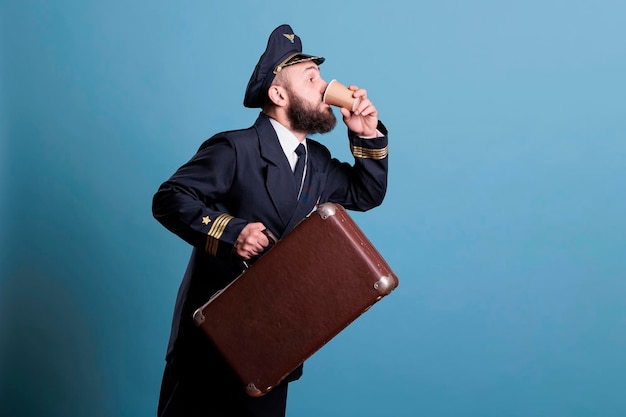 Capitán de avión con uniforme profesional de aerolínea que llega tarde, lleva maleta, bebe café para llevar. Piloto con equipaje en el aeropuerto, aviador con equipaje con taza de papel de té, vista lateral