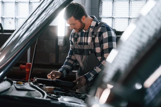 Capô do carro é aberto Usando tablet Homem de uniforme está trabalhando no salão de automóveis