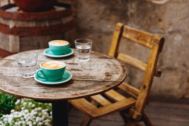 Foto cappuccino de aroma matinal com espuma de leite exuberante por cima em duas xícaras de cerâmica turquesa com copos de água em uma mesa redonda de madeira