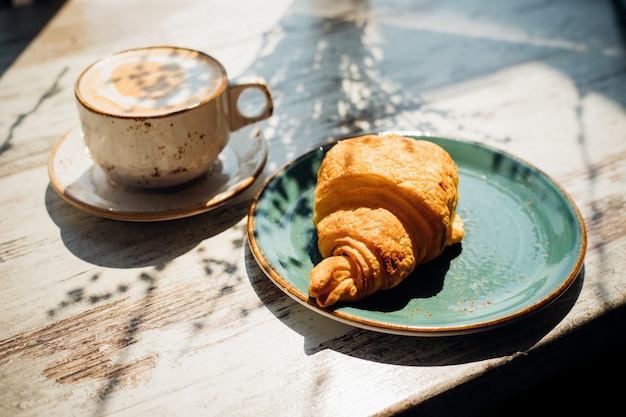 Cappuccino e croissant estão na mesa do café. O sol da manhã cai sobre a mesa, lindas sombras aparecem.