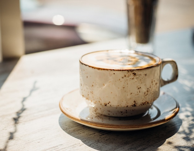 Cappuccino in einer schönen Keramiktasse steht auf dem Tisch in einem Café. Morgensonne fällt auf den Tisch, schöne Schatten erscheinen. Leckeres aromatisches Frühstück.