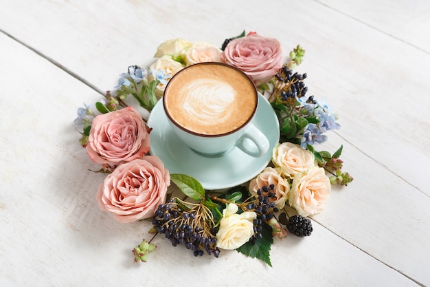 Cappuccino und blumen zusammensetzung. blaue kaffeetasse mit cremigem schaum, frischen und getrockneten blumen kreisen am weißen holztisch. heiße getränke, saisonales angebotskonzept