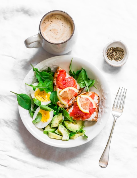 Cappuccino und eine Schüssel mit grünem Salat, Avocado, gekochtem Ei und Sandwiches mit Roggenbrot, Frischkäse und Lachs auf hellem Hintergrund, Draufsicht Köstlicher und gesunder Frühstücksbrunch