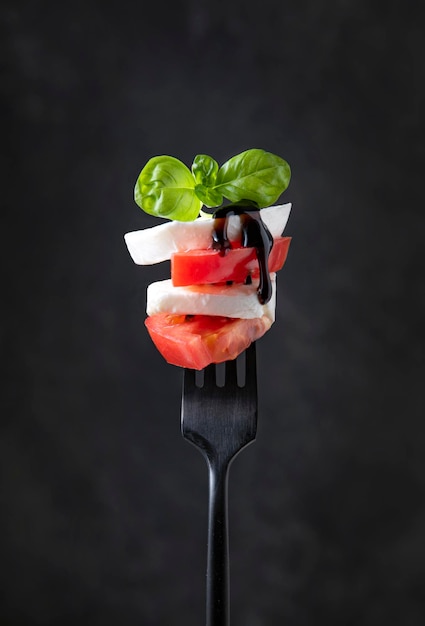 Caprese-Salat-Mozzarella schneidet frische Tomaten und frische Basilikumblätter mit Balsamico-Sauce auf einer schwarzen Gabel auf dunklem Hintergrund