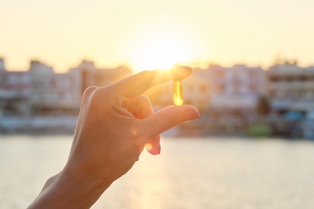 Cápsula de gel amarillo vitamina d omega-3 en mano de mujer, fondo de mar de sol.