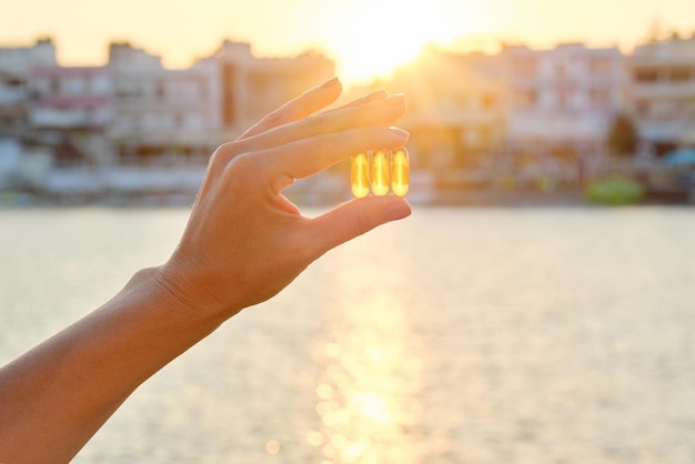 Cápsula de gel amarillo vitamina d omega-3 en mano de mujer, fondo de mar de sol.