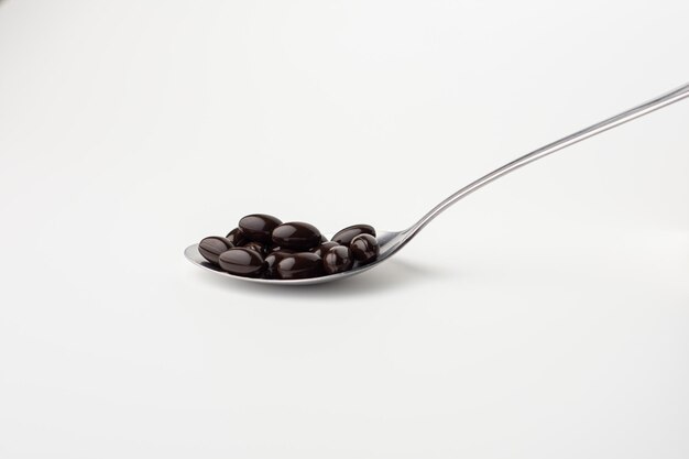 Cápsulas de gelatina blanda negra en una cuchara de acero inoxidable sobre un fondo blanco. complementos alimenticios vitamínicos