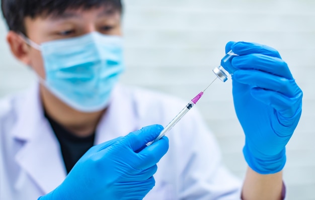 Captura aproximada da agulha da seringa sugando a vacina coronavírus covid19 do frasco de vidro para injeção por cientista masculino profissional asiático em jaleco branco usando máscara facial em fundo desfocado.